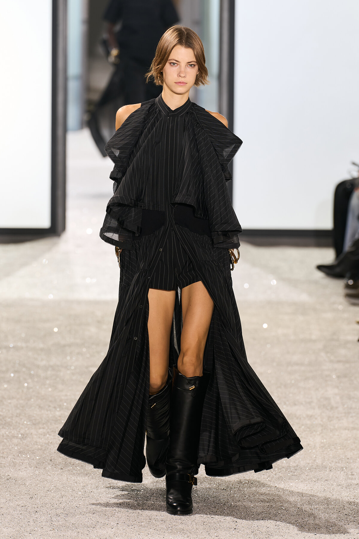 Model on a runway wearing a black pinstripe ensemble with voluminous layered cape sleeves and knee-high boots.