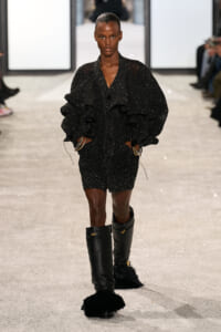 Model walks the runway in a black speckled suit with voluminous ruffled sleeves, matching shorts, and tall fur-trimmed boots on a glittery catwalk.