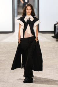 Model walking a runway in a black and cream sculptural top over a sheer blouse, paired with wide black trousers and gold bracelets.