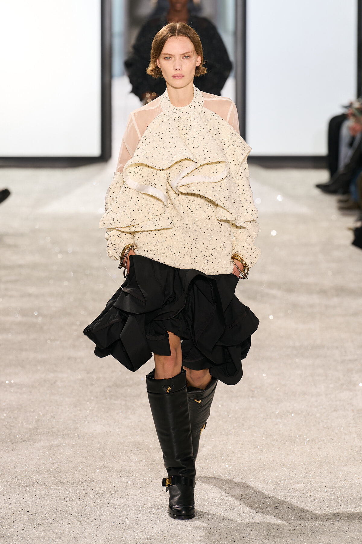 Runway model wearing a cream speckled ruffled knit top with sheer sleeves and a voluminous black layered skirt, paired with tall black knee boots.