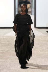Runway model wearing an all-black look: striped top, flowing sheer cape, loose black pants, and gold bracelets.
