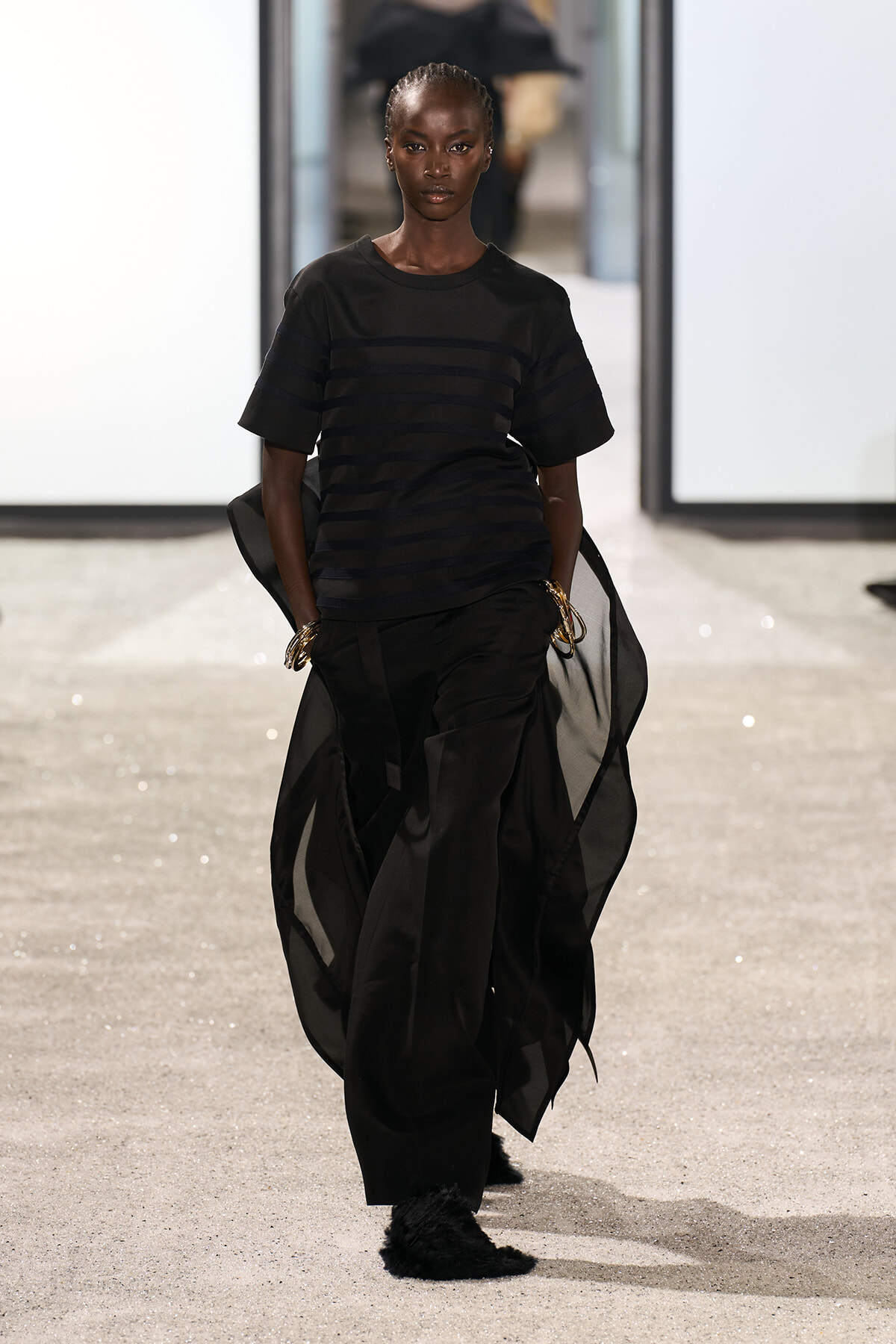 Runway model wearing an all-black look: striped top, flowing sheer cape, loose black pants, and gold bracelets.