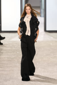 Model walks the runway in a black sculptural jumpsuit with a beige ruffled neck scarf and bracelets.
