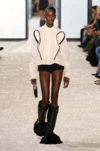 Model on a runway wearing a cream oversized shirt with black piping, black shorts, tall black fur-trimmed boots, and gold bracelets.