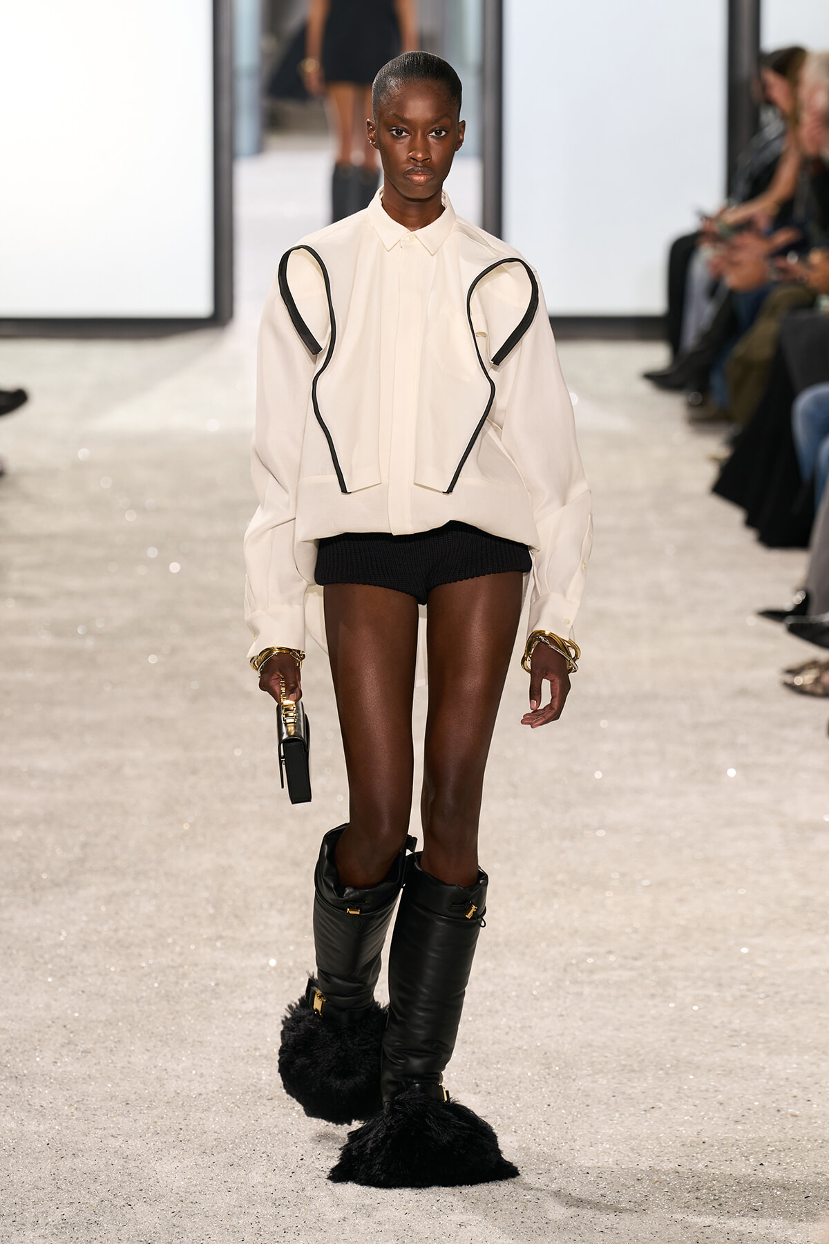 Model on a runway wearing a cream oversized shirt with black piping, black shorts, tall black fur-trimmed boots, and gold bracelets.