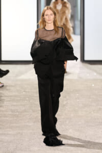 Female model walking a runway in a black, oversized suit with a sheer mesh top and dramatic folded cape-like jacket.