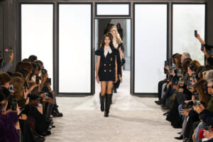 Models walk a glittery runway flanked by seated audiences filming with smartphones; front model wears a navy double-breasted coat-dress with knee-high boots.