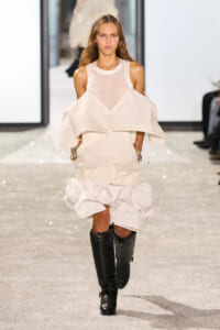 Model walking a runway in a white, layered dress with an off-the-shoulder cape detail and tall black knee-high boots.