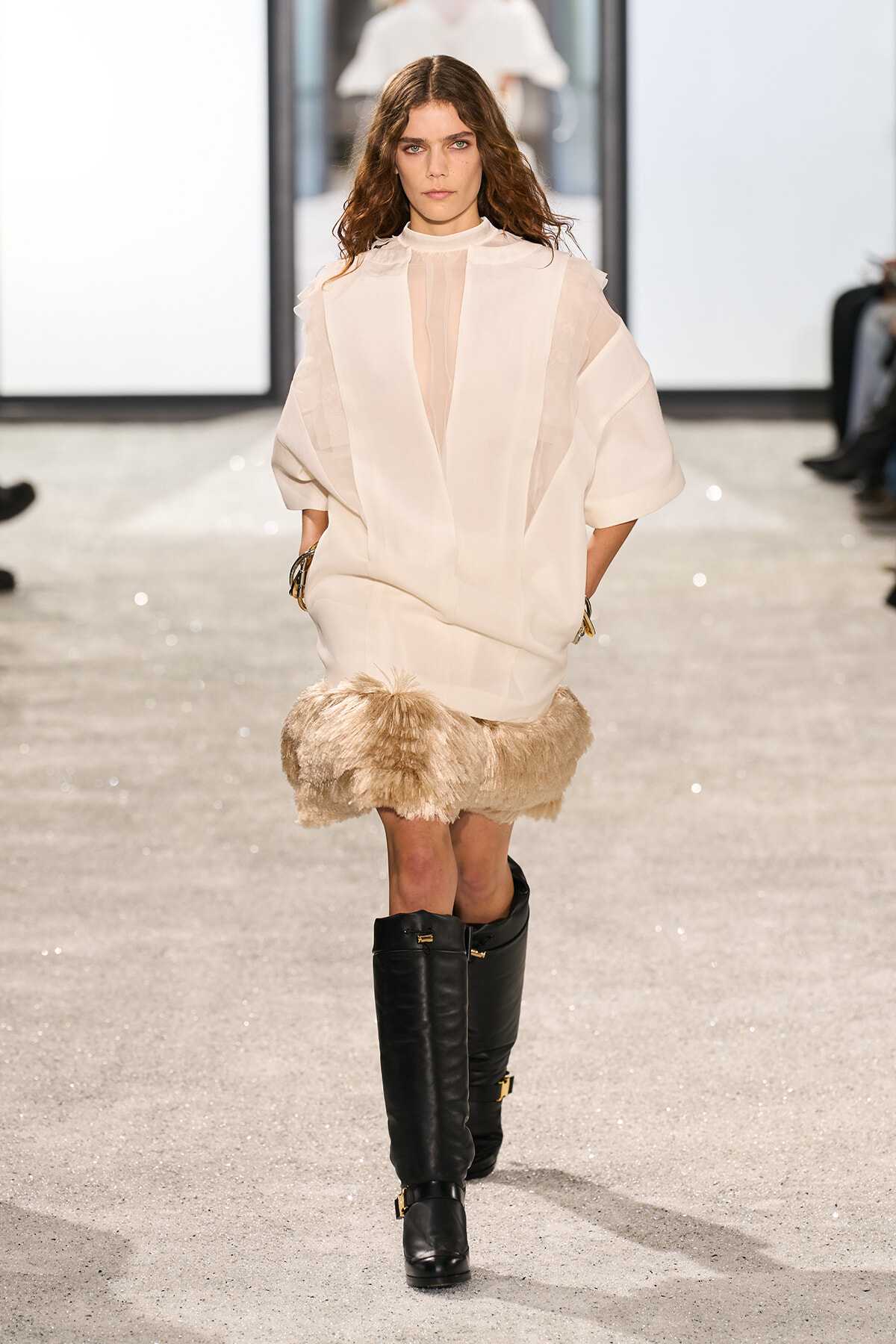 Female model walking a fashion runway in an oversized ivory tunic with sheer panels and a fur-trimmed beige mini skirt, paired with tall black leather boots.