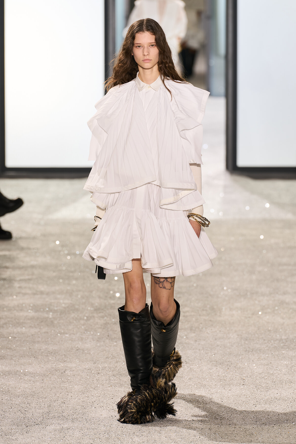 Model walking a runway in an all-white layered outfit with a ruffled cape and black fur-trimmed boots.