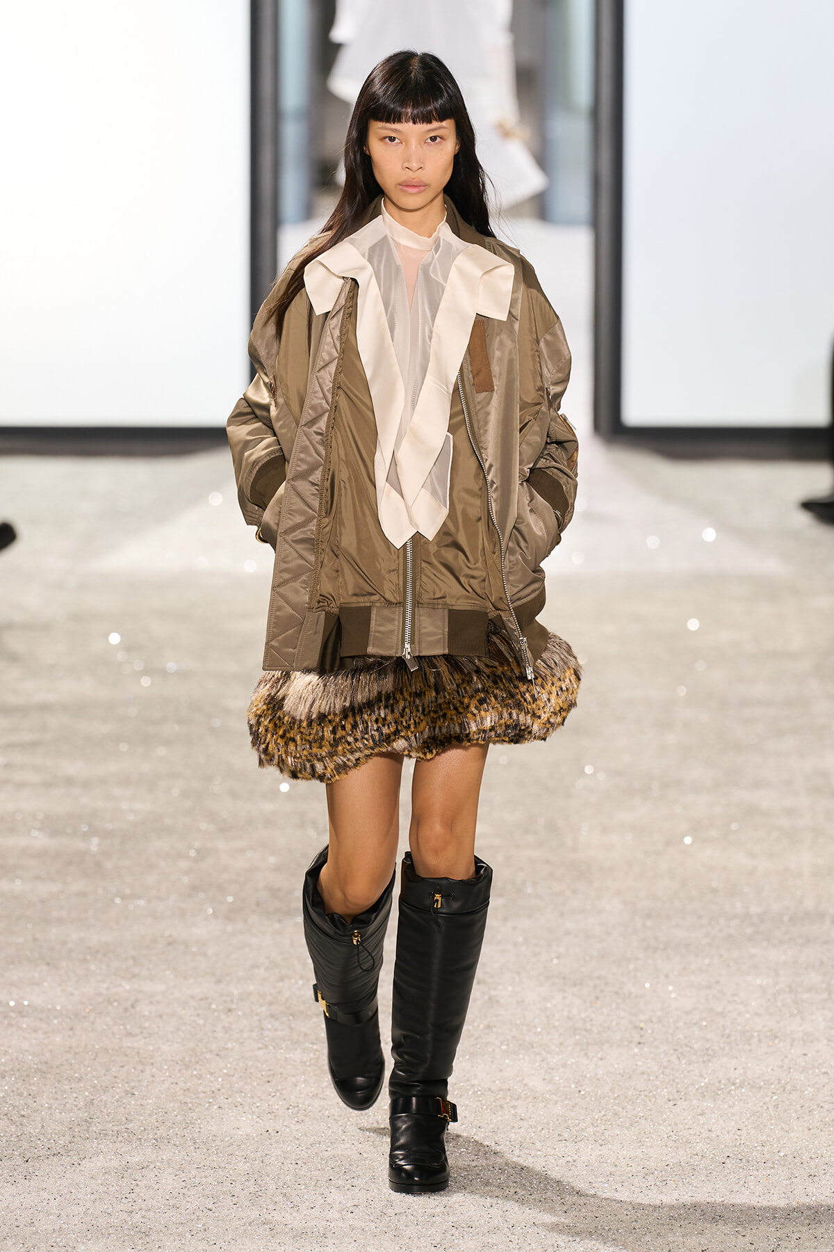 Model on a runway wearing a tan oversized jacket with white front panels, a fur-trimmed mini skirt, and black knee-high boots.