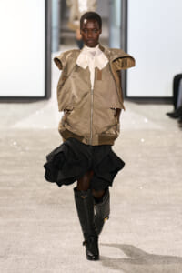 Model walks the runway wearing a tan oversized jacket with a white scarf-like collar, paired with a voluminous black skirt and knee-high boots.