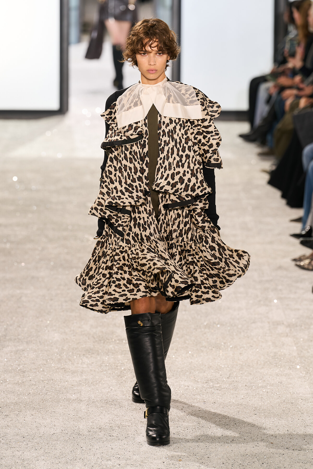 Model walks a fashion show in a cream leopard-print, tiered ruffled dress with a large pale collar and black knee-high boots.