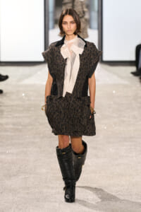 Model walking a runway in a dark leopard-print cape dress with oversized shoulders, cream neck scarf, and black knee-high boots.