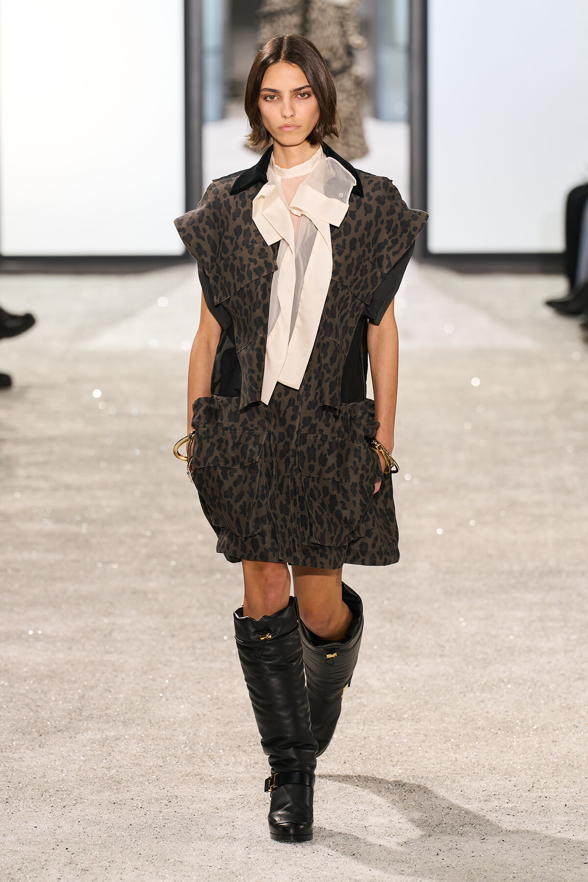 Model walking a runway in a dark leopard-print cape dress with oversized shoulders, cream neck scarf, and black knee-high boots.
