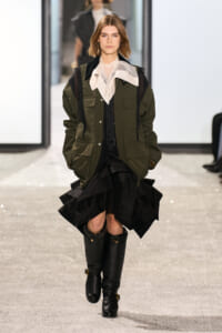 Model walking runway in an olive-green utility jacket over a white blouse, with a black layered skirt and knee-high boots.