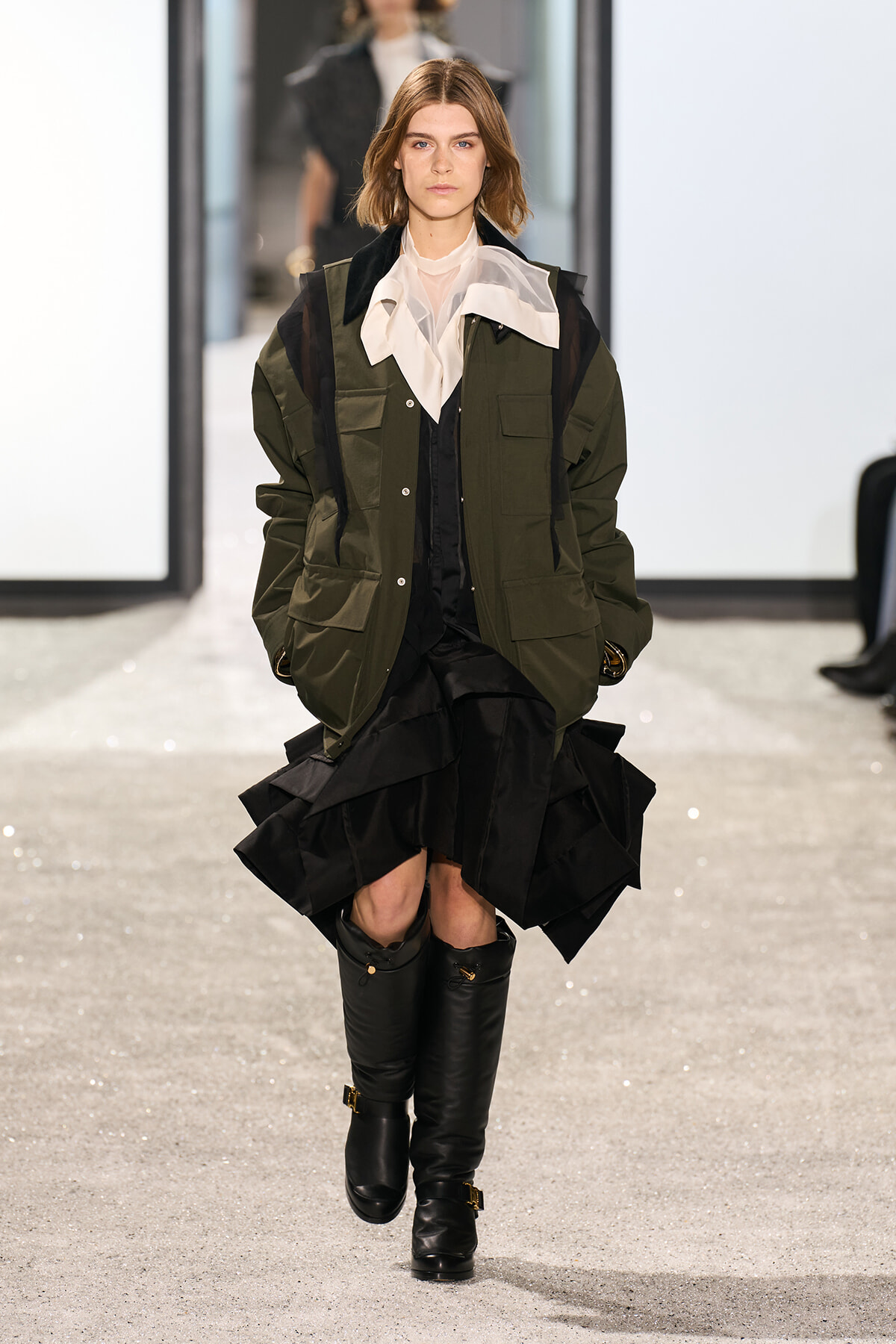 Model walking runway in an olive-green utility jacket over a white blouse, with a black layered skirt and knee-high boots.
