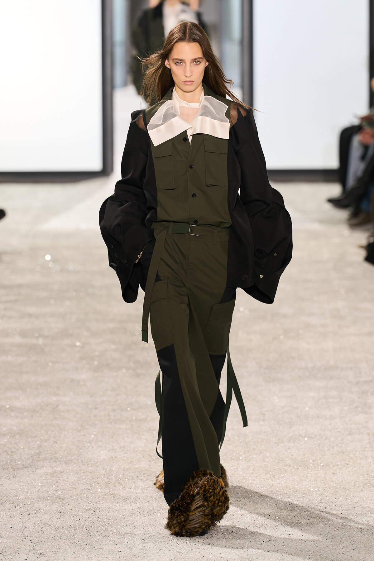 Model walking runway in olive utility jumpsuit with black oversized sleeves and dramatic white collar; fur-covered footwear visible at the front.