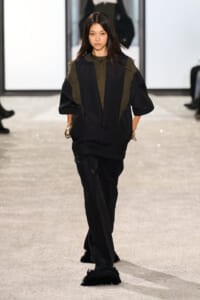 Female model walks a runway in a loose black top with olive panel accents and black wide-leg pants, wearing furry black shoes and gold bangles on both wrists