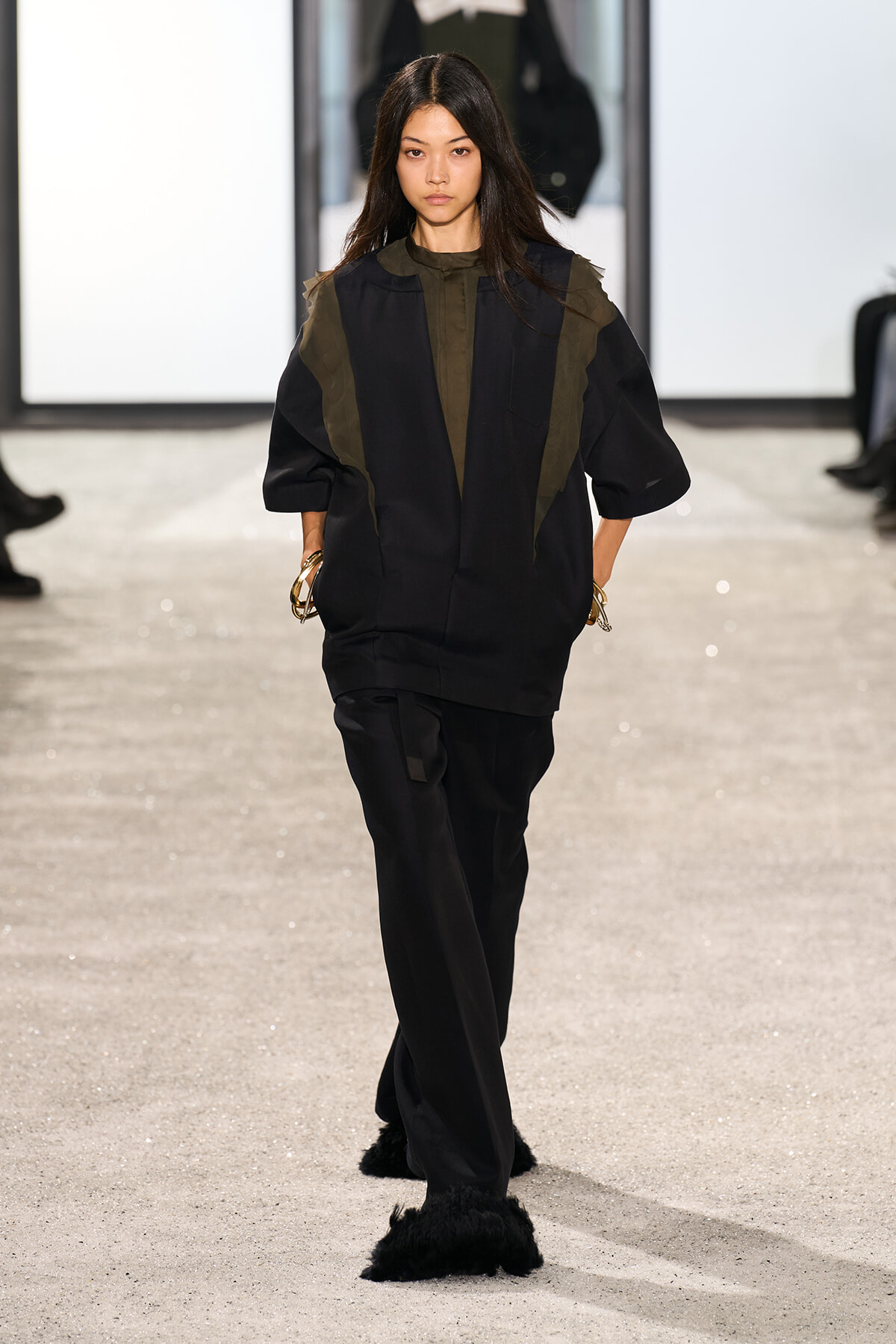 Female model walks a runway in a loose black top with olive panel accents and black wide-leg pants, wearing furry black shoes and gold bangles on both wrists