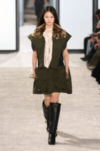 Model walking a runway in an olive green utility dress with large pockets, a fur-collared cape, and knee-high black boots.