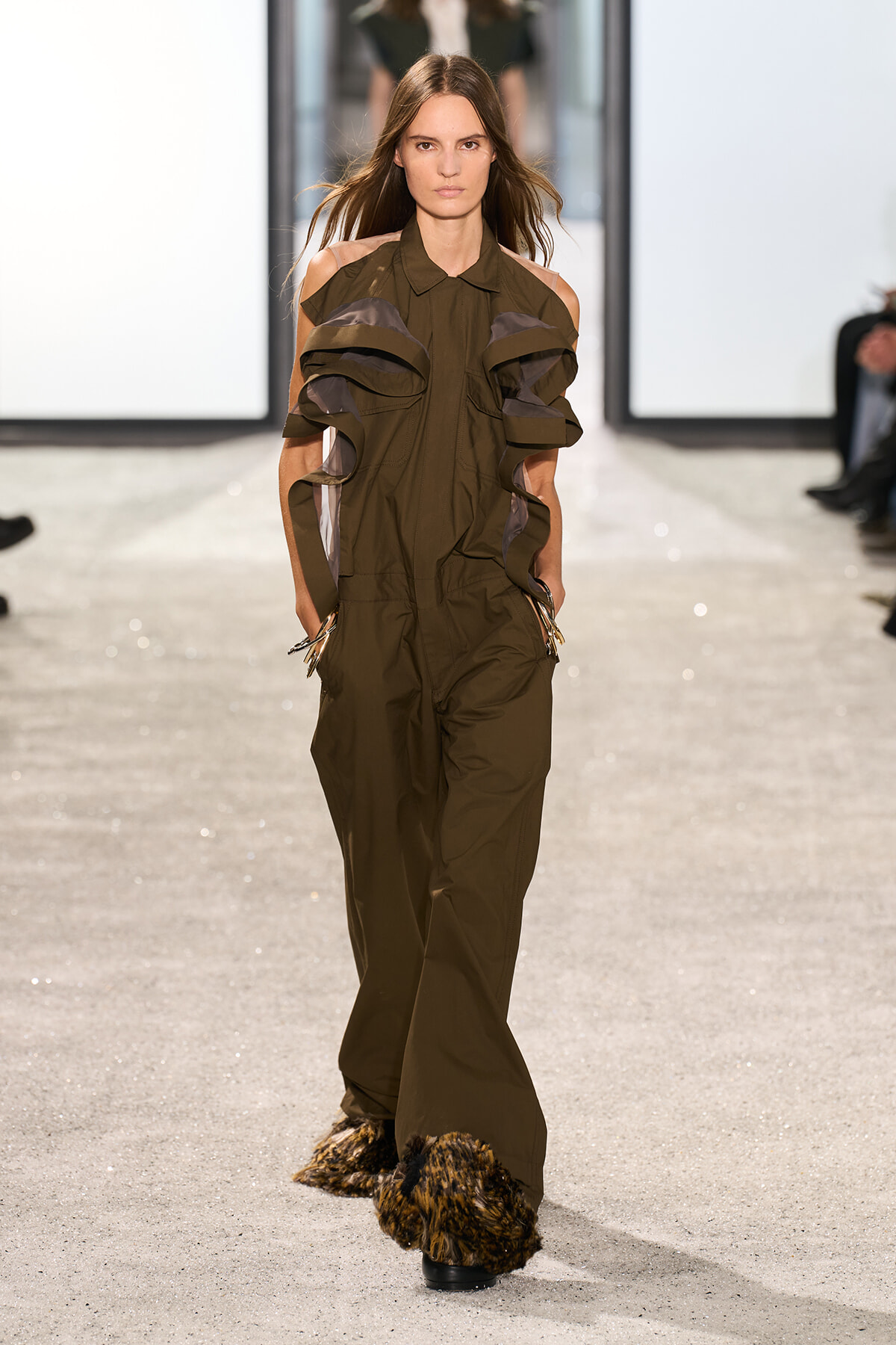 Model walks a runway in an olive-green jumpsuit with dramatic sculpted ruffles along the sleeves and fur-trimmed pant cuffs.