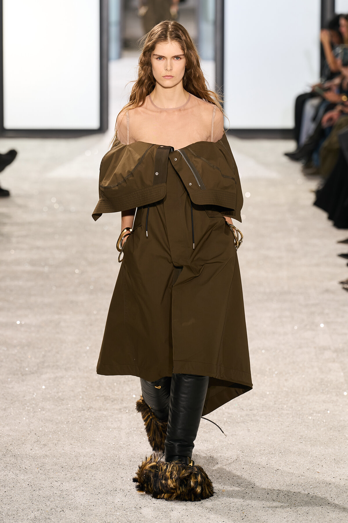 Model walking a runway in an oversized olive-brown coat draped off the shoulders over a sheer pink top; fur-trimmed boots visible at the ankles.