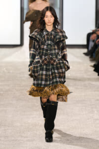Female model on a runway wearing an oversized plaid coat/dress with a fur fringe hem and black knee-high boots.