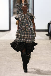 Runway model wearing a plaid, layered top with puff sleeves and a voluminous black ruffled skirt, paired with knee-high socks and buckled boots.