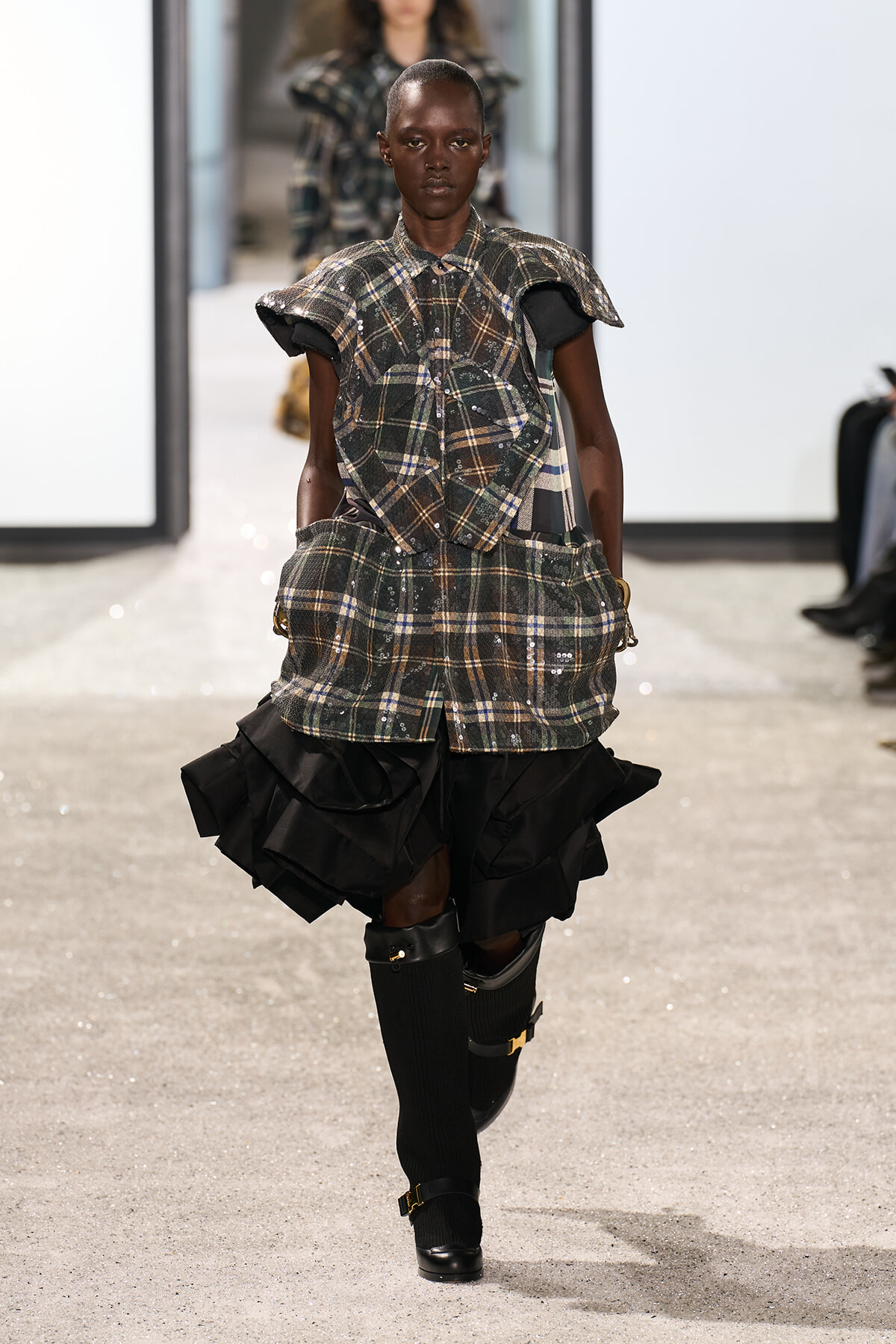 Runway model wearing a plaid, layered top with puff sleeves and a voluminous black ruffled skirt, paired with knee-high socks and buckled boots.