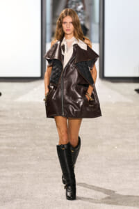Model on a runway wearing an oversized dark brown leather coat with quilted black panels and a white shirt underneath, paired with knee-high black boots.