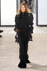 Model walking a fashion show in a black sheer blouse with a high ruffled neck, an oversized dark denim jacket, belt, and black wide-leg pants with fuzzy black slippers.