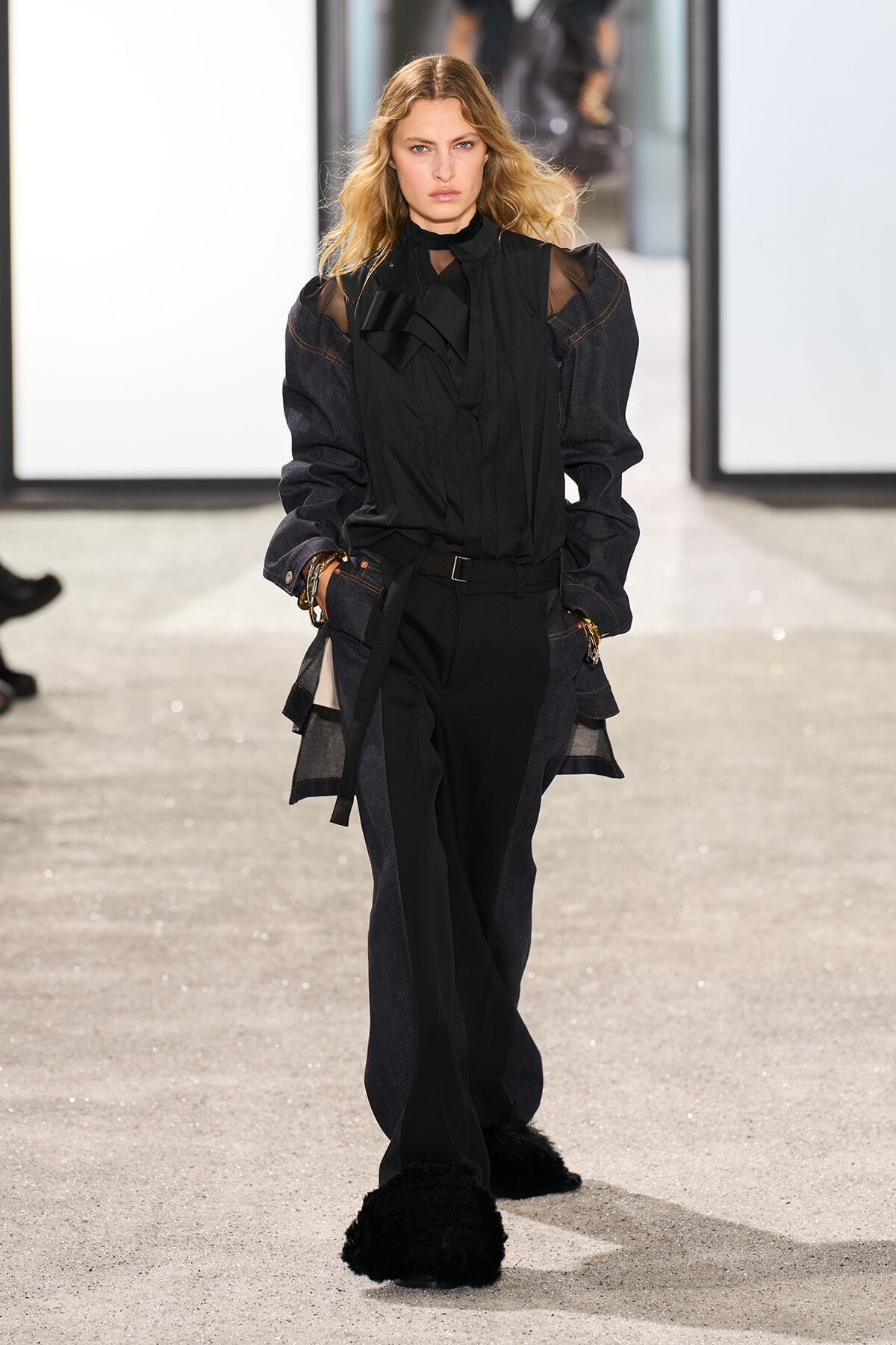 Model walking a fashion show in a black sheer blouse with a high ruffled neck, an oversized dark denim jacket, belt, and black wide-leg pants with fuzzy black slippers.