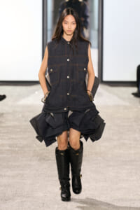 Model walking a runway in a sleeveless dark denim vest over a voluminous layered denim skirt and black knee-high boots.