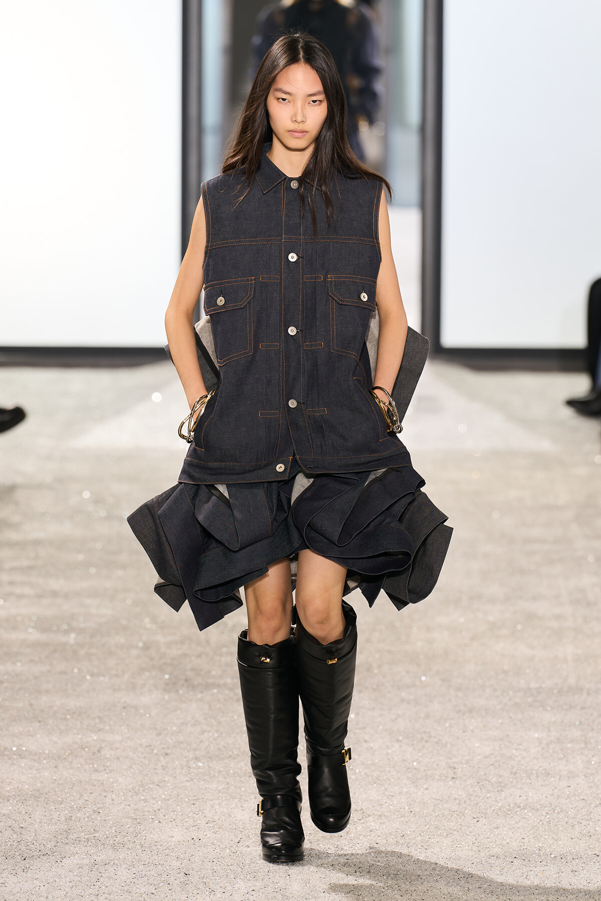 Model walking a runway in a sleeveless dark denim vest over a voluminous layered denim skirt and black knee-high boots.