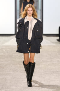 Model walking runway in oversized dark denim jacket with white blouse and tall black boots.