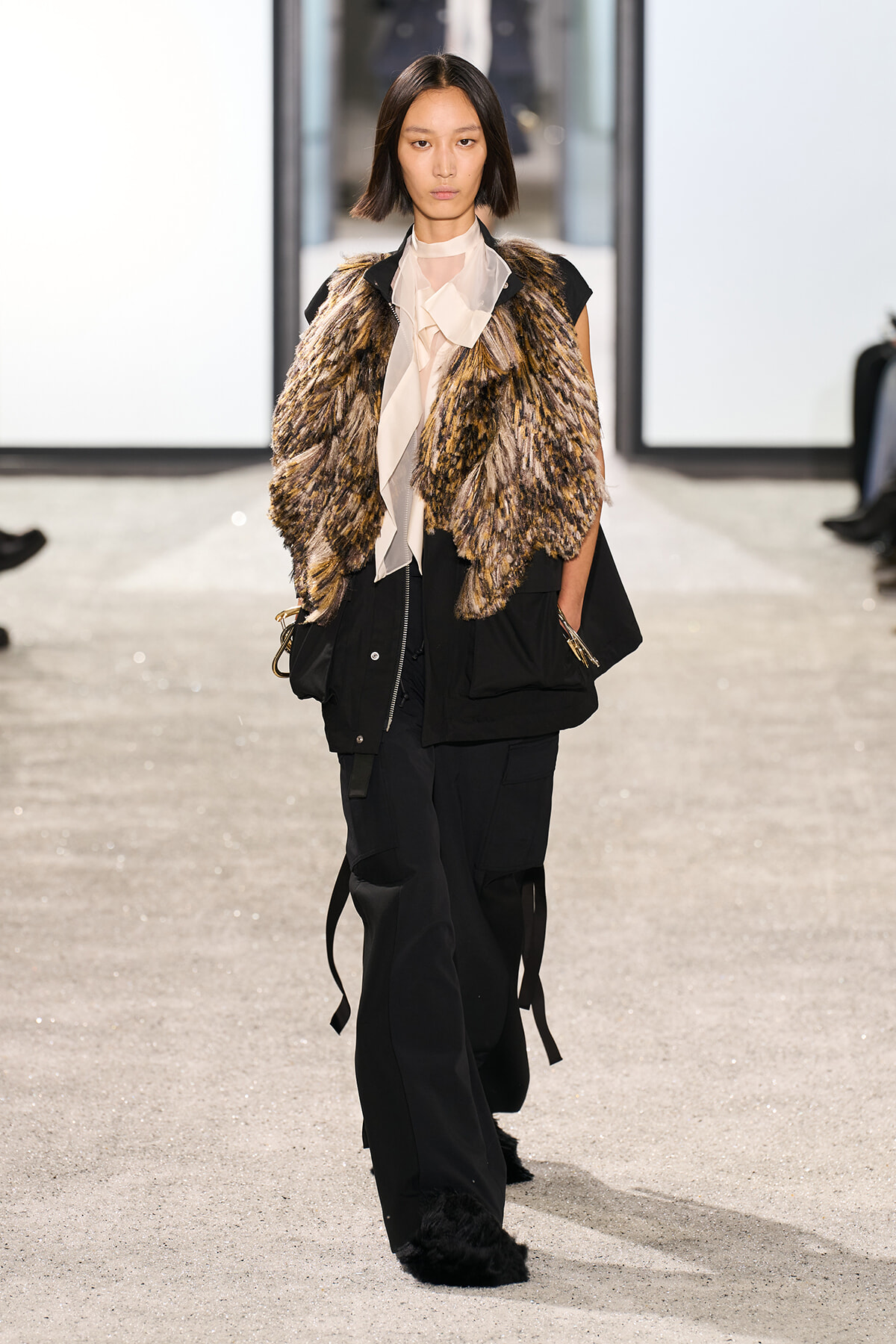 Model walking a fashion runway in a fur-textured vest over a pale blouse and oversized black pants.