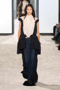 Model walking a fashion runway in a navy sleeveless coat over a cream blouse, paired with wide-leg jeans and fur-trimmed hems.
