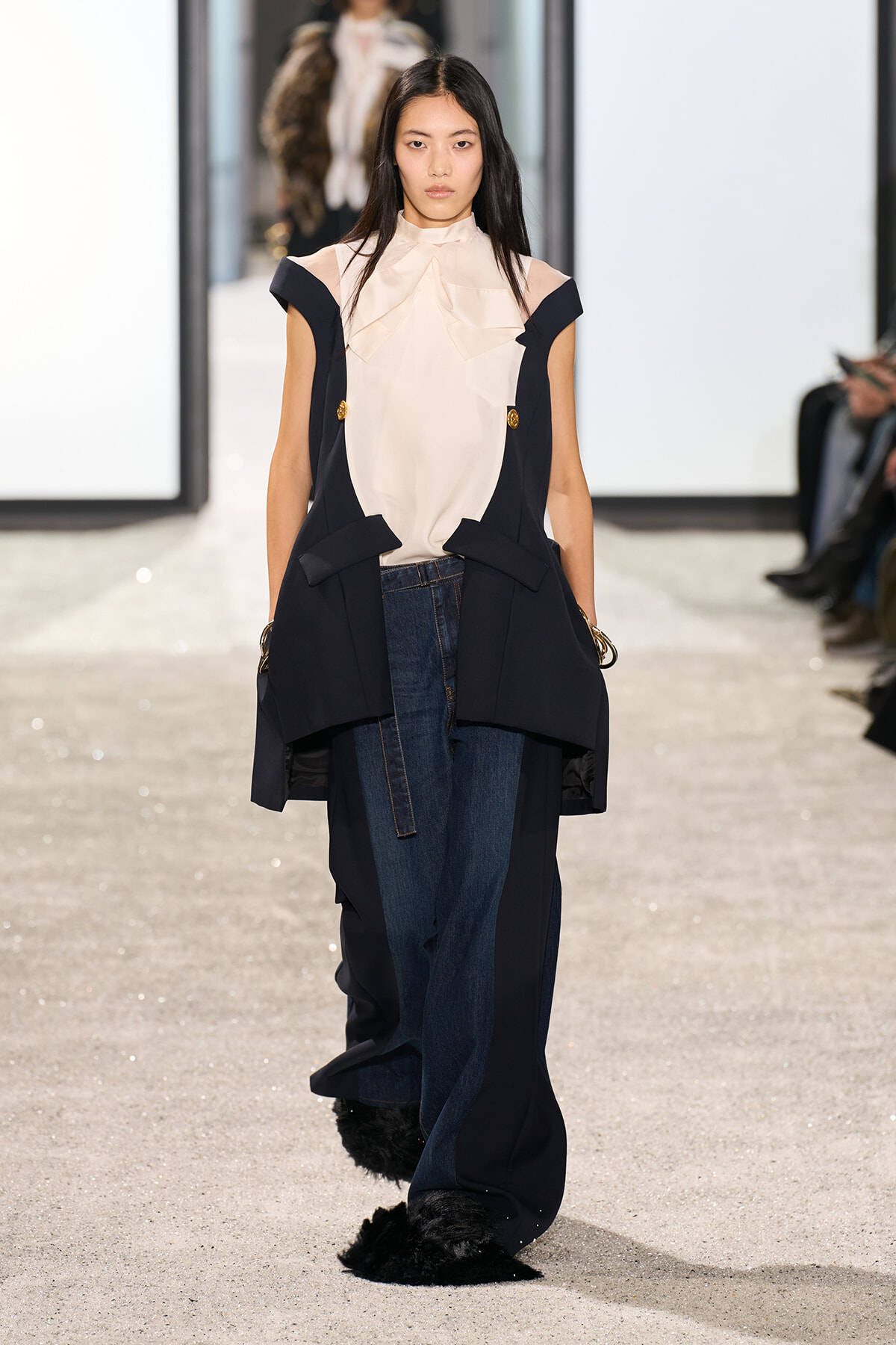 Model walking a fashion runway in a navy sleeveless coat over a cream blouse, paired with wide-leg jeans and fur-trimmed hems.