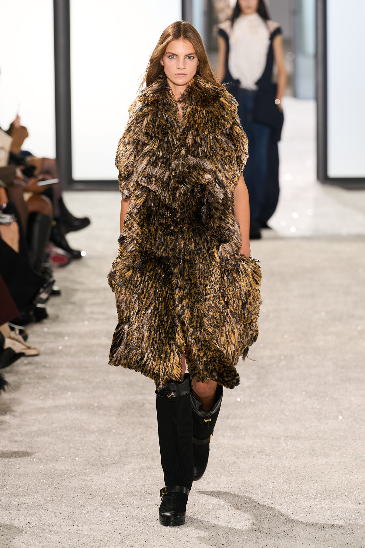 Model walking runway in a voluminous brown shaggy fur coat over black knee-high boots.
