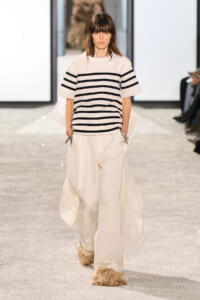 Model walking a fashion runway in a cream striped short-sleeve top and wide cream trousers, with fur-covered shoes.