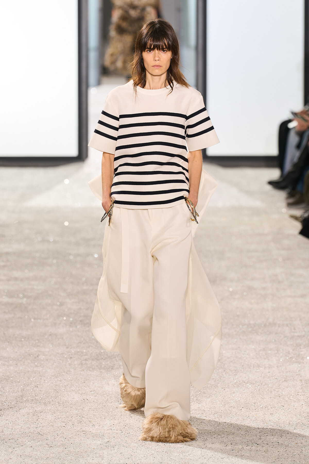 Model walking a fashion runway in a cream striped short-sleeve top and wide cream trousers, with fur-covered shoes.