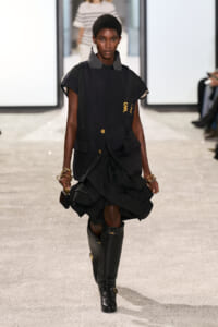 Model walking runway in a structured black sleeveless vest over a dark dress with a ruffled hem, tall black boots, and gold jewelry.