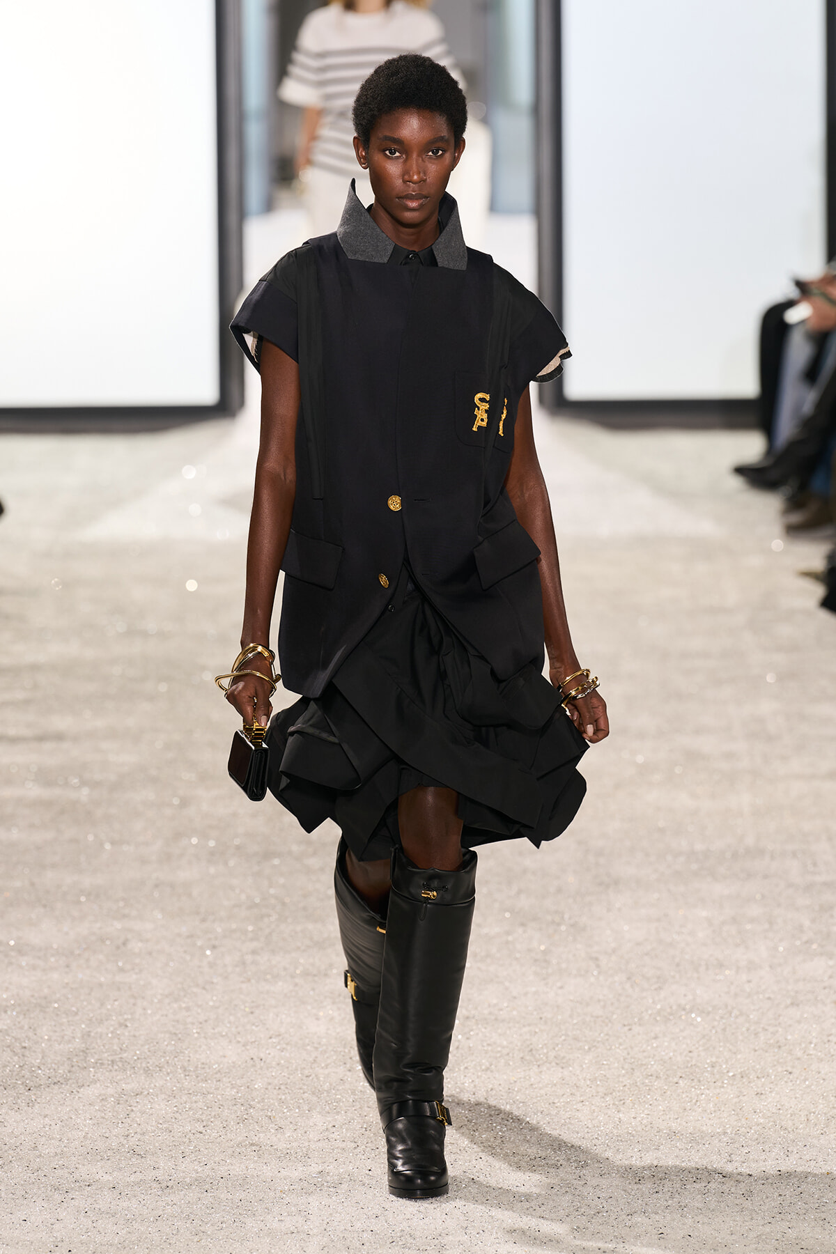 Model walking runway in a structured black sleeveless vest over a dark dress with a ruffled hem, tall black boots, and gold jewelry.