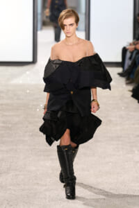 Model walks the runway in a dramatic black, multi-layered dress with oversized ruffles and a sheer nude top, paired with knee-high black boots.