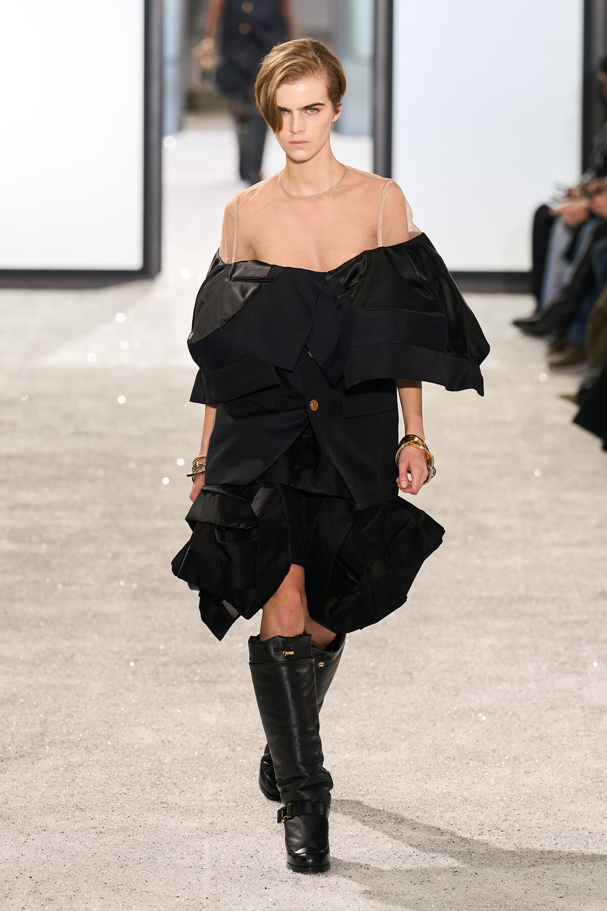 Model walks the runway in a dramatic black, multi-layered dress with oversized ruffles and a sheer nude top, paired with knee-high black boots.