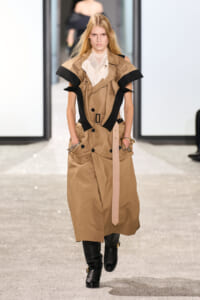 Runway model wearing a tan oversized trench with black panel accents, a belted waist, and black boots.