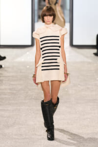 Model walks the runway in a cream dress with black horizontal stripes and side pockets, paired with knee-high black boots at a fashion show.