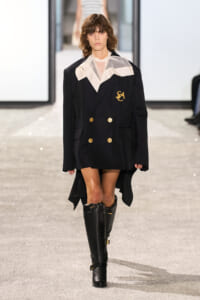 Model walking a runway in a navy double-breasted blazer with gold buttons, white collar, and tall black boots.