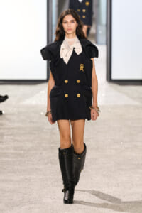 Model walking the runway in a black double-breasted mini dress with gold buttons, oversized shoulders, and a beige bow tie at the neck, paired with tall black boots and bracelets.